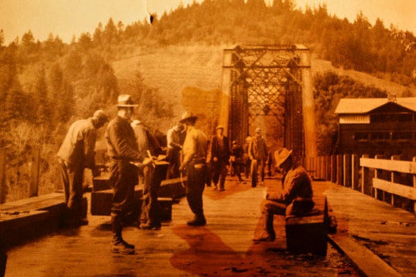 Sepia-toned vintage photo of people gathered on wooden bridge with forested hills and rustic structures in background