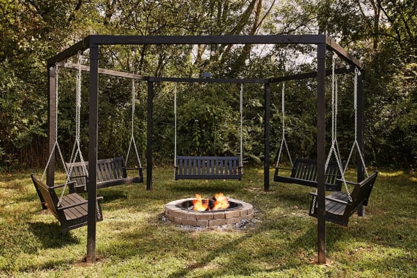 Black metal swing set with multiple swings surrounding a stone fire pit with active flames in a wooded backyard.